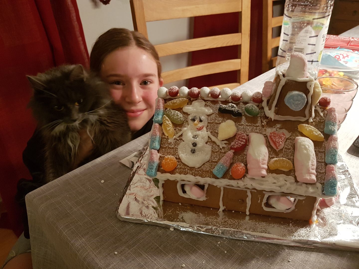 A girl holding the grey cat next to a gingerbread house decorated with sweets and an icing snowman.